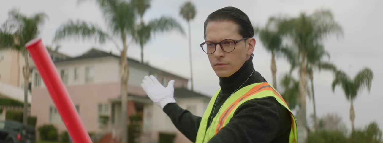 A young man wearing reflective clothing is directing traffic near a residence in a tropical area
