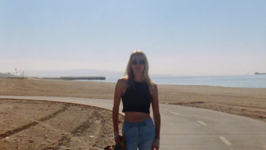 A teenage girl wearing sunglasses with long blond hair is standing on a road through the beach holding her skateboard