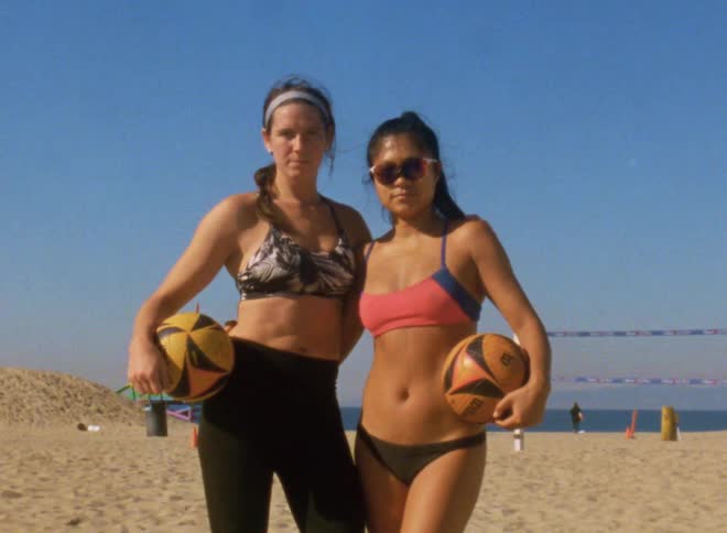 Two friends holding volleyballs in front of a net in the beach hugh each other looking at the camera