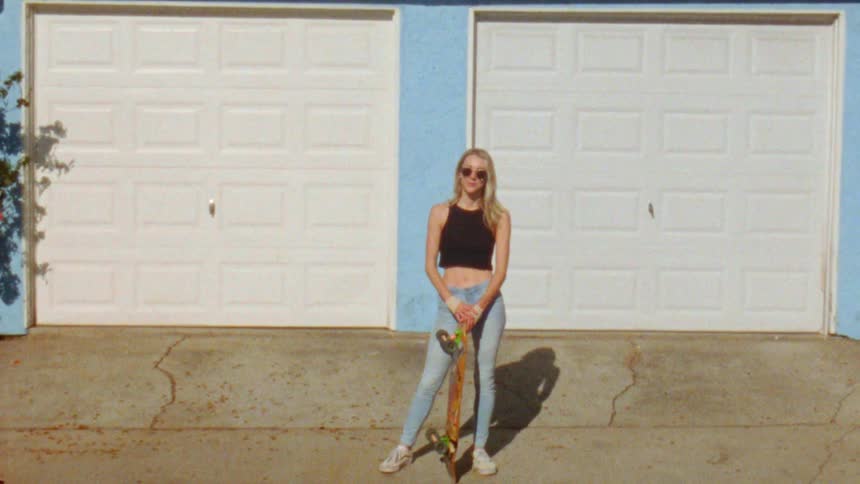 A young blonde woman is standing in a driveway and posing with a skateboard resting against her hand
