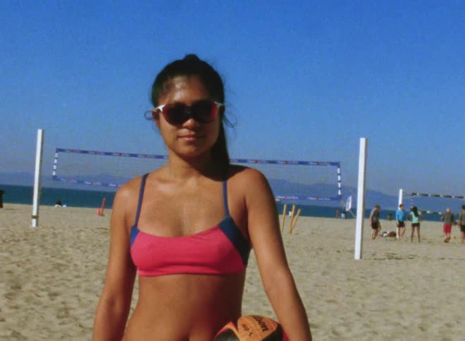 A woman in a swimsuit looks into the camera while posing with a volleyball on a beach during the day