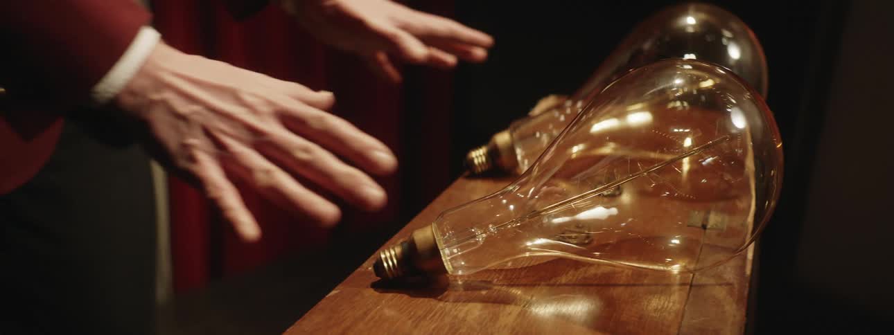 Large light bulbs sit on a wooden table and someone comes up and puts their hands on them