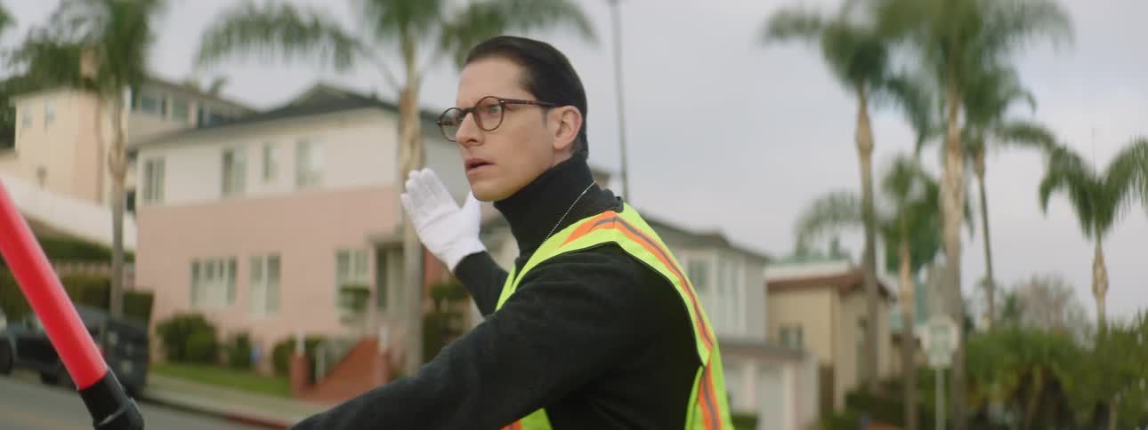 A man wearing white gloves and a yellow vest is holding a red baton as he stand in the street directing traffic