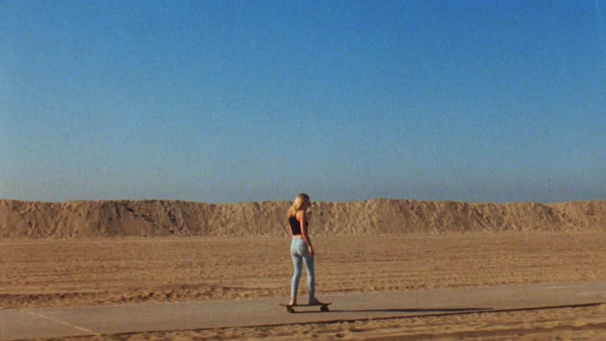 The sun shines upon a teenage girl with long blond hair as she rides her skateboard on a road through the beach
