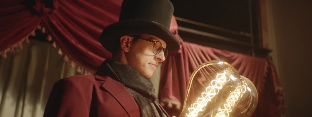 A man wearing a tall black hat and black scarf holds two large light bulbs in his hands