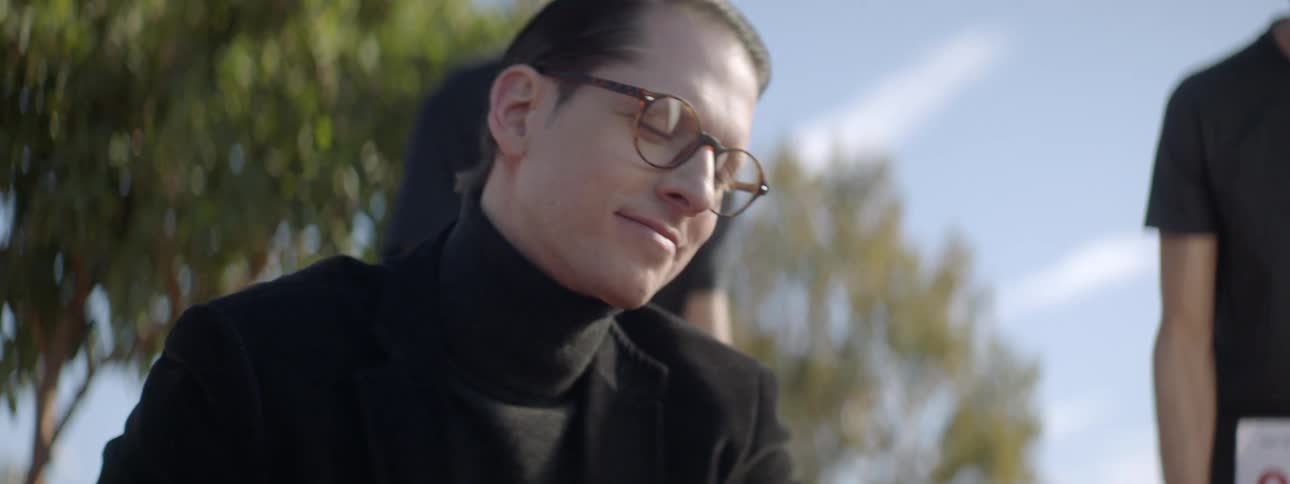 Man with glasses and a black turtleneck leans down as two boys join him examining something on the ground outside