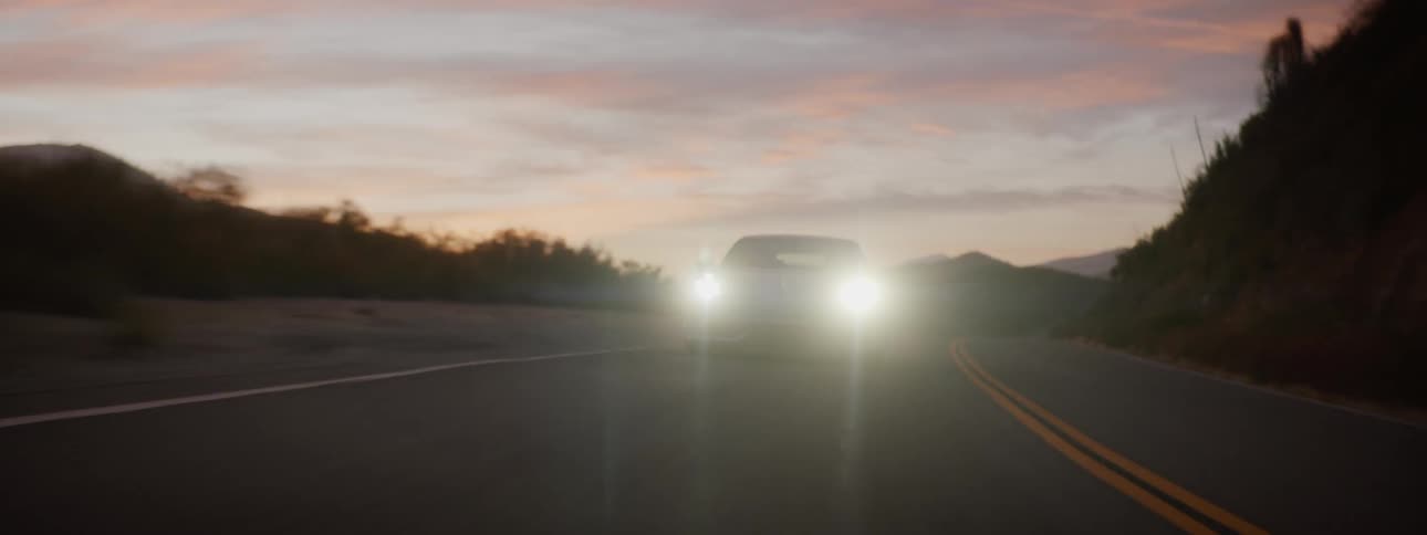 A white sports car with the headlights on is traveling on a desert road with mountains in the distance