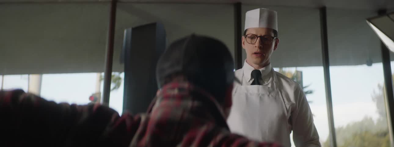 A server wearing a white apron and hat smiles at a customer who aggressively throws a napkin down and walks away