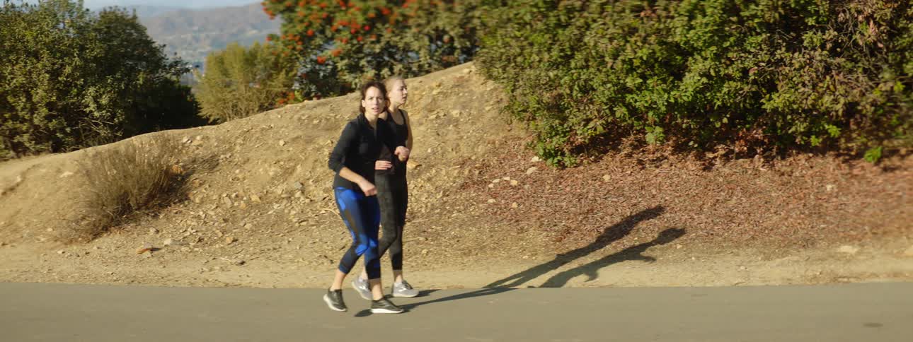 Two teenage girls are jogging up a hill and they stop to look to the side and then keep running