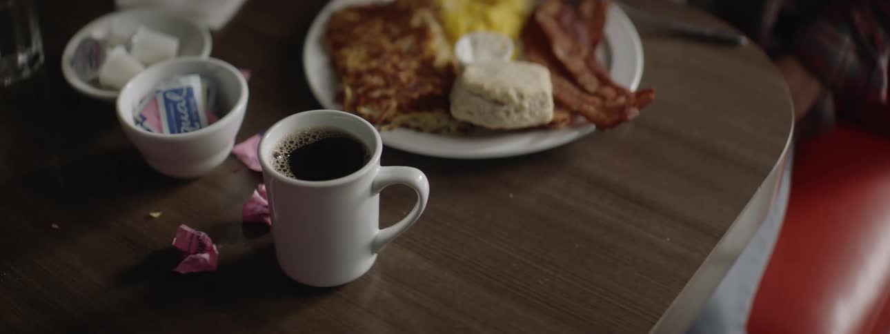 A waiter places a cup of coffee next to a breakfast plate and another person touches them with their hand to see if they are hot