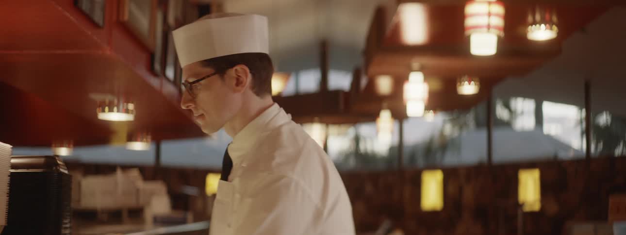A man wearing all white and a white cap walks and then pours something in a cup
