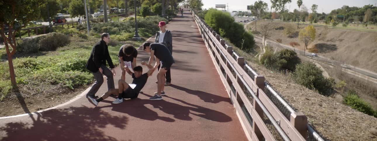 Two teenage runners are standing near another boy lying on the ground and an older man standing near them holding a clipboard as another man comes to aid the boy on the ground and pushes his chest and the boy wakes up as the others help him stand