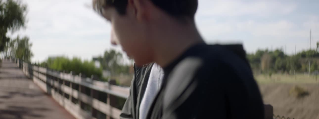An elderly man is standing near the railing of a walkway as a teenage boy walks near him he seems annoyed and motions with his hand as he walks away