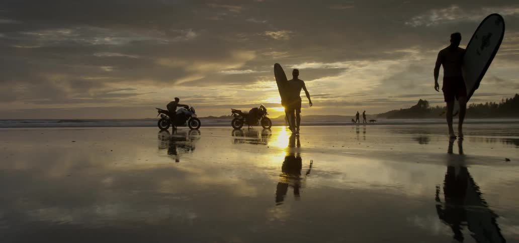 People carrying surfboards towards the water's edge with two motorcycles on the beach
