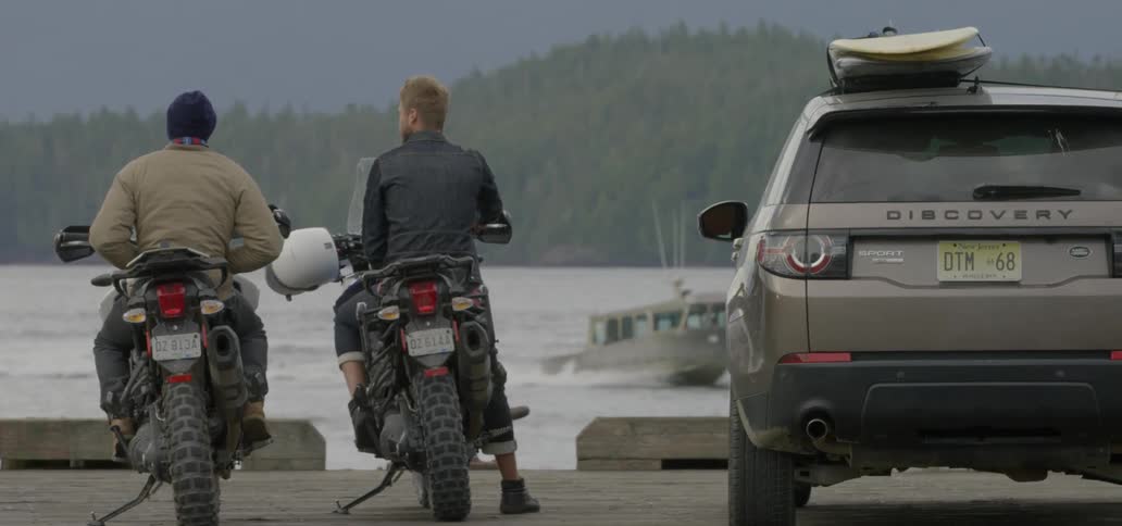 Two men sit on motorcycles near the water