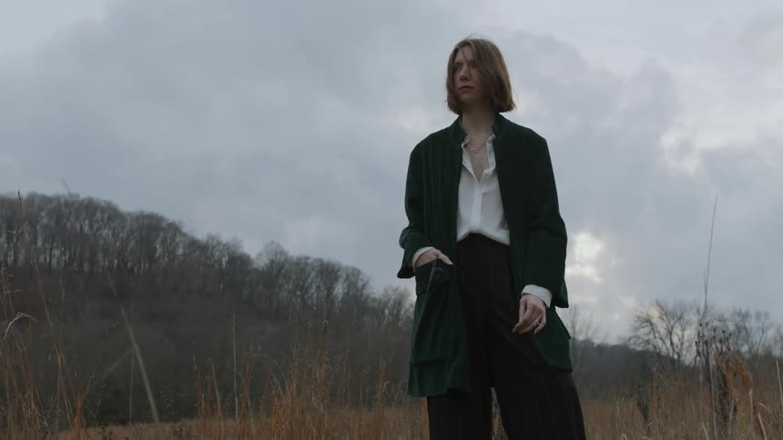 A woman wearing a white shirt with black pants and a jacket is standing outside in an open field with her hand in her pocket and there are bare trees in the distance