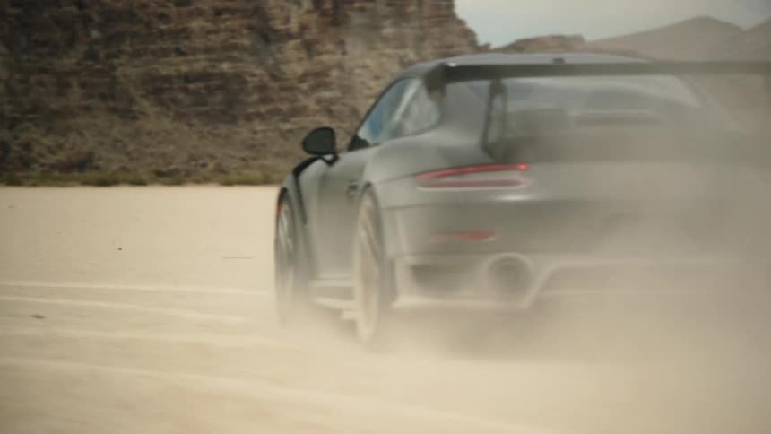 A dark sports car with a rear spoiler leaves a trail of dust as it speeds across the desert and there are mountains in the distance