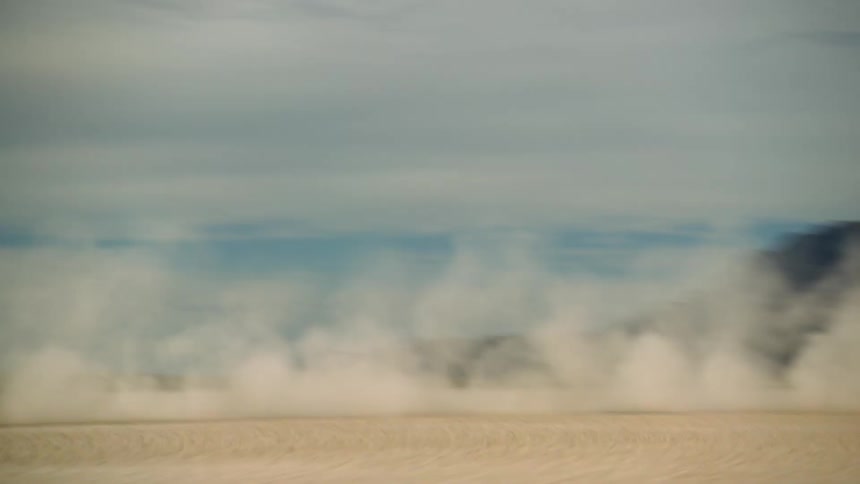 A dark sports car leaves a trail of dust as it speeds across the desert and there are mountains in the distance under a cloudy sky