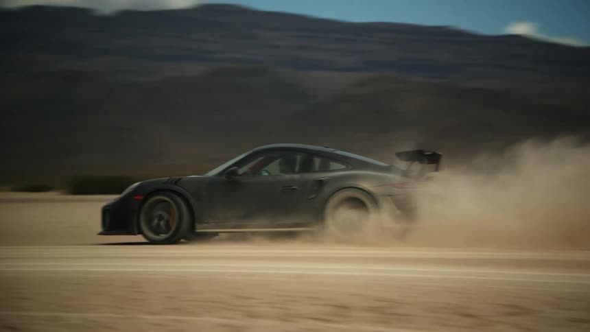 A dark sports car leaves a trail of dust as it speeds across the desert and there are mountains in the distance