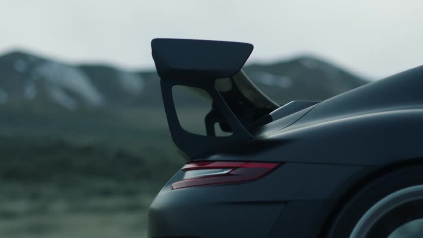 A dark sports car with a rear spoiler travels on a road with mountains in the distance