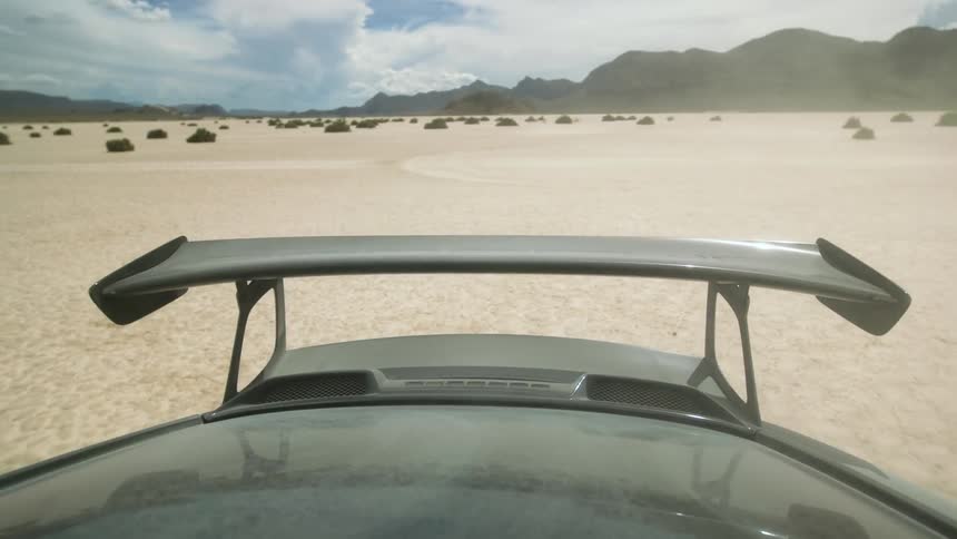 A cars spoiler is in view as the car drives around a sandy deserted area wildly
