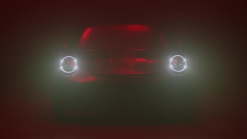 An automobile glowing red is sitting in the darkness as rings of light shine around the headlights then the headlights come on and emit a bright ray of light in front of the car