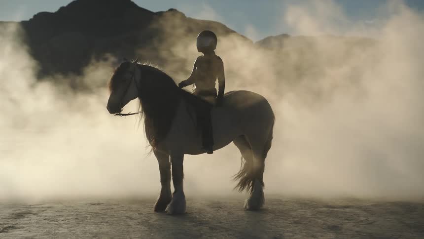 A woman sits on a horse in front of a mountain range as dust blows over them