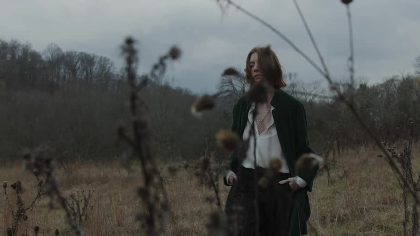 Woman with brown hair black coat white blouse and pants walk in the field on a cloudy day