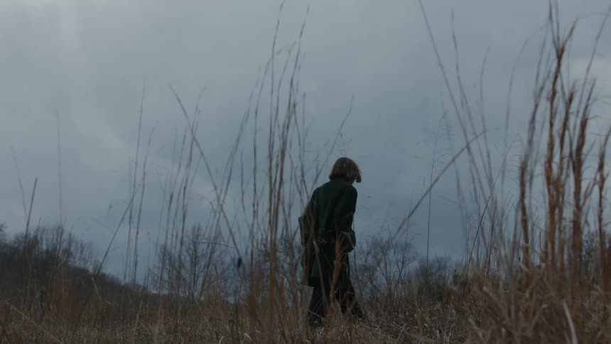 A woman wearing a long black coat walks slowly up a hill against a grey sky