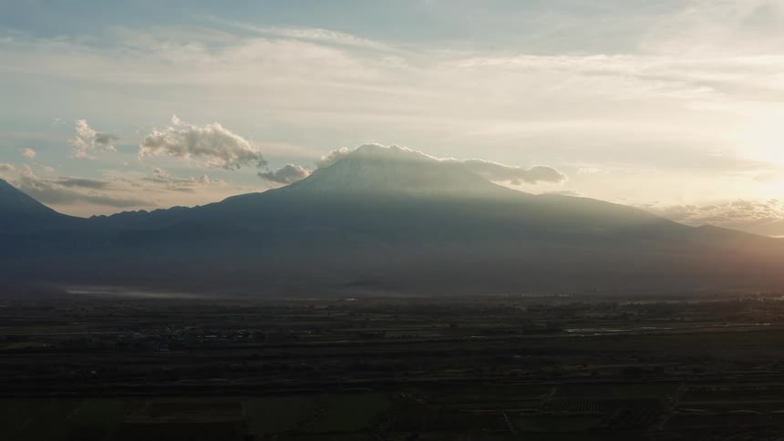 A mountain stands high in the clouds across the river from a neighborhood as the sun shines brightly on the horizon