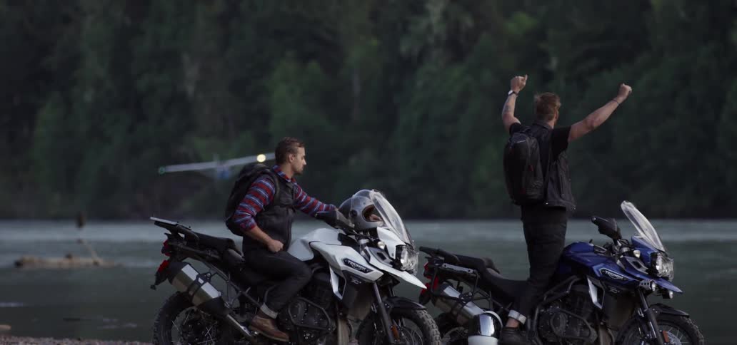 Two men with motorcycles raise their arms in signal to a plane landing on water near them