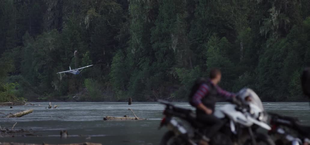 A plane flying very low over a river as two motorcyclists watch