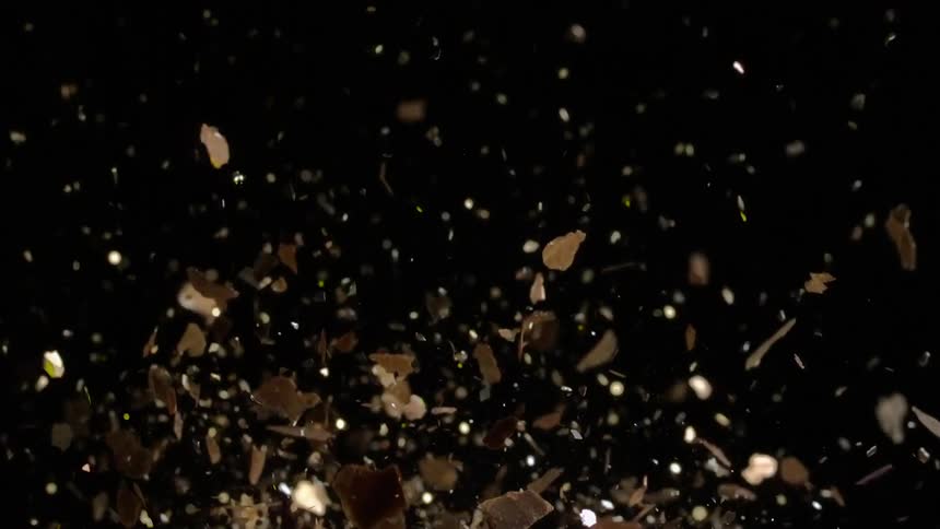 An object is shattering and shards of broken pieces shine and glitter as they fly into the darkness