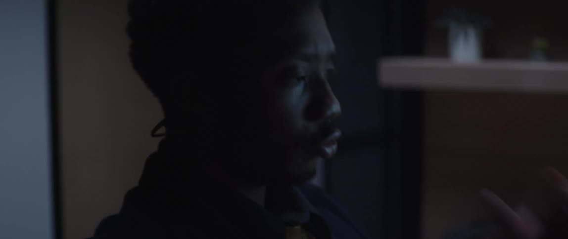 A man is sitting at a desk pushing buttons on a phone under a computer monitor and he leans back in his chair