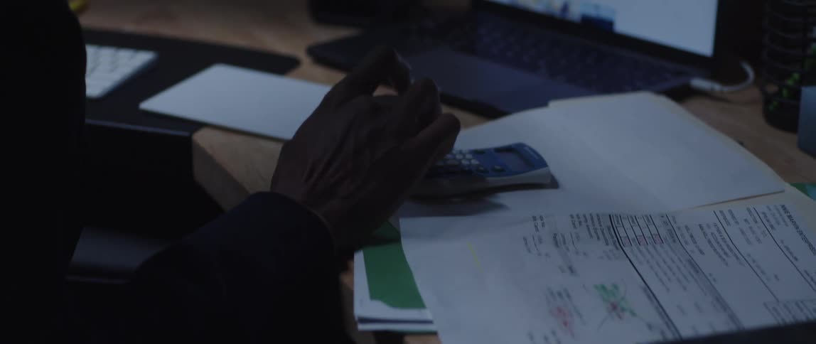A man is sitting at a desk with a computer and a calculator as he goes through paperwork and makes calculations