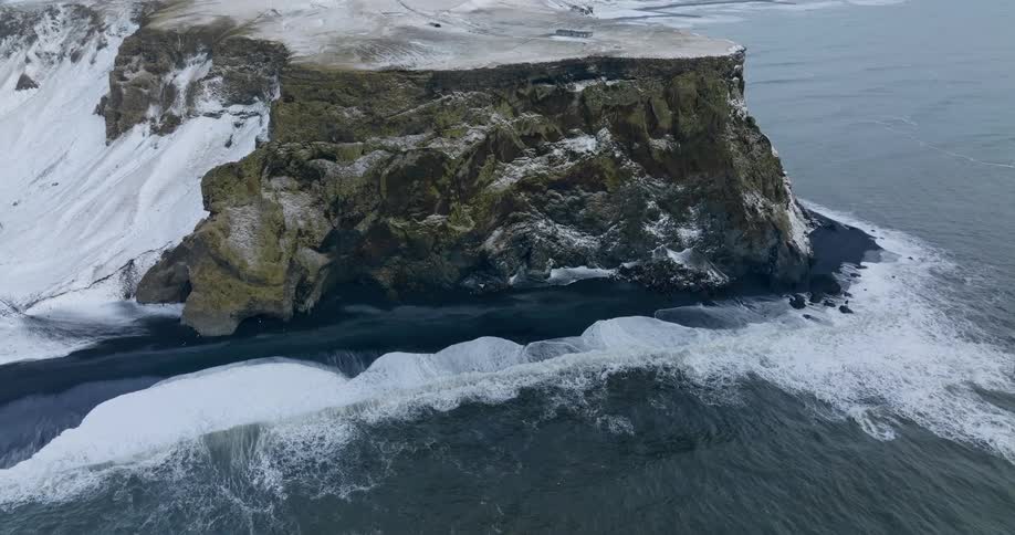 Aerial view of ocean waves breaking against the black beach near snowy cliff