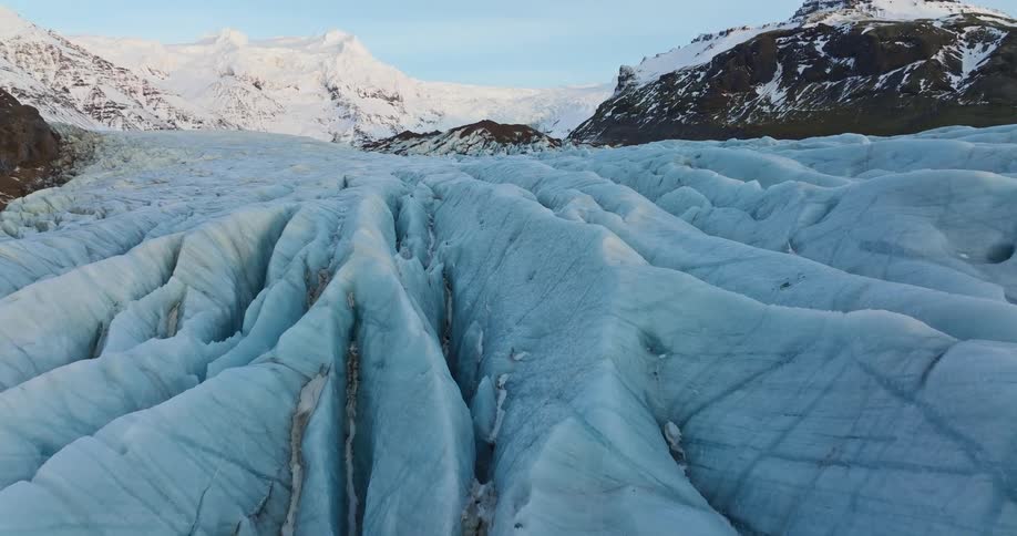 Frozen snow covered ridges stand across the ground and there are snow covered mountains in the distance under a clear sky