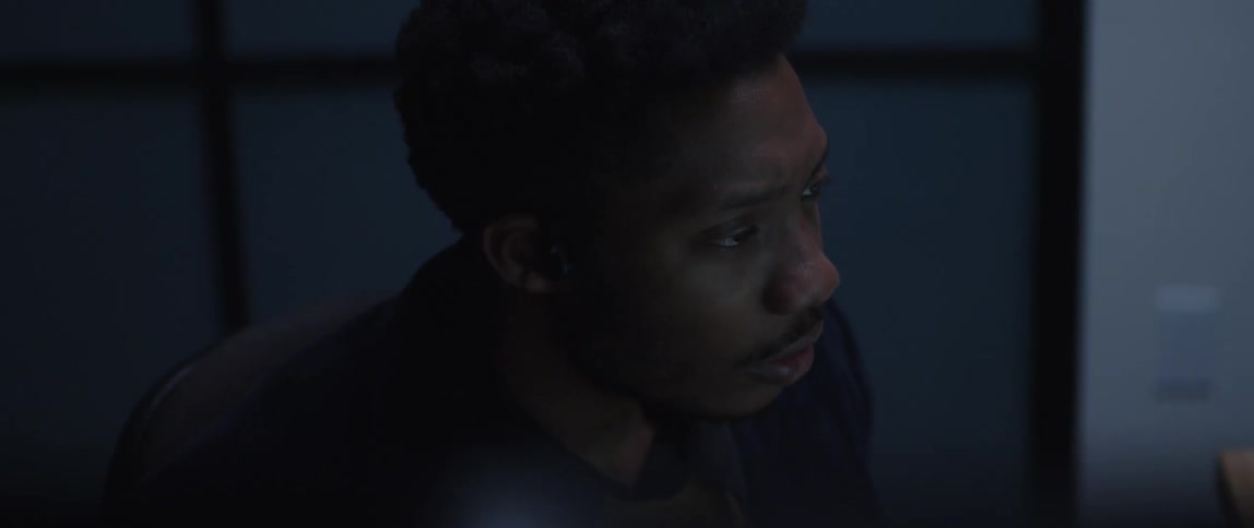 A man is sitting at a desk holding a pen and looking at a computer screen and he hurries to write something down
