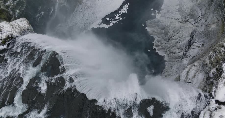 Water falls over a snowy mountain ledge down to the river below