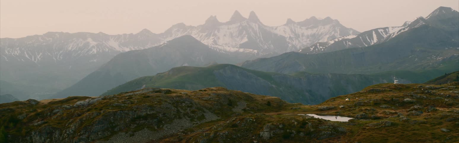 A frozen lake and a small building are on top of a mountain and there are mountains in the distance with peaks high in a hazy sky