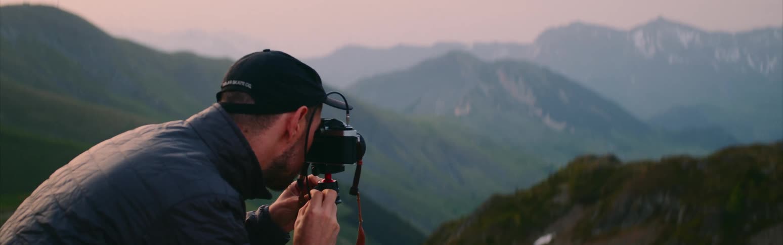 Photographer wearing black cap and coat take photos of mountain range with camera on tripod