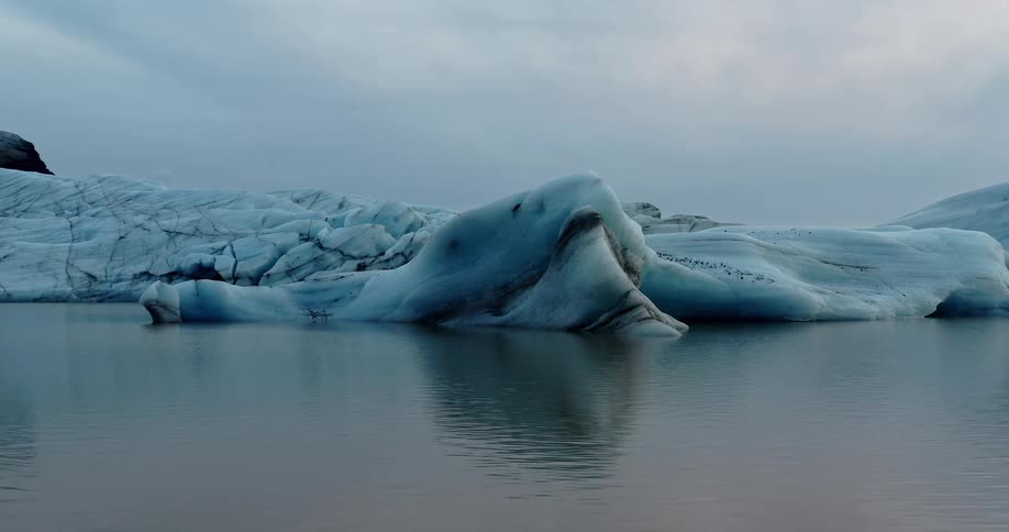 Water near shore reflects iceberg under an overcast sky