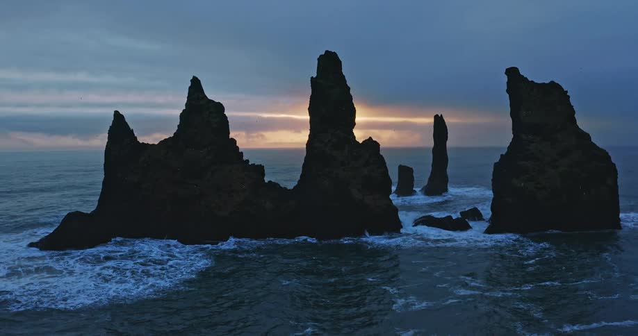Sun sits over the horizon in red sky casting rock outcroppings surrounded by white waves partly into silhouettes