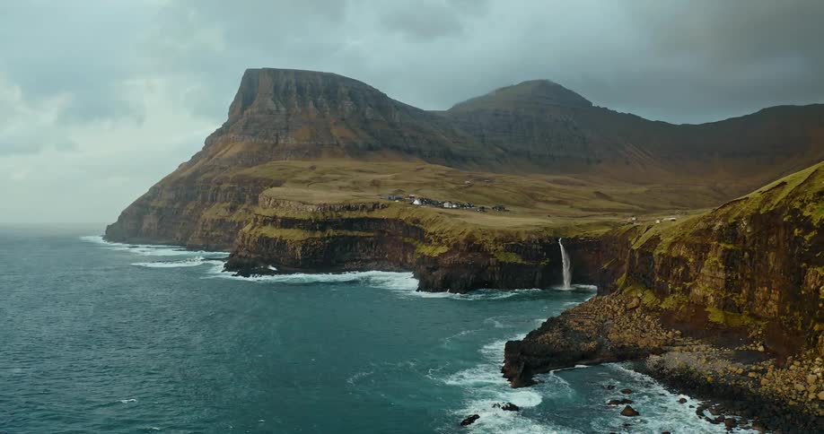 Waves crash against the island cliffs along the coastline as mountains stretch high into a cloudy hazy sky