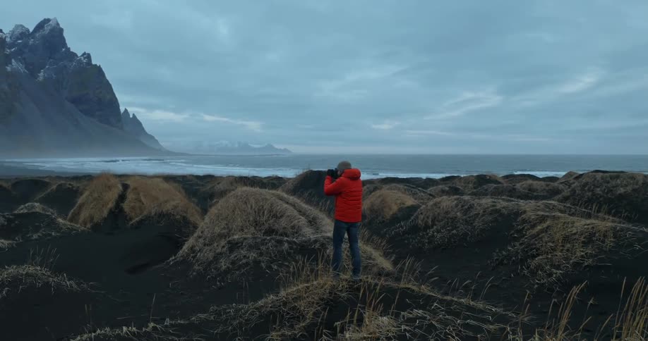Male photographer in red coat and jeans takes photos of high mountain near the ocean on a cloudy day