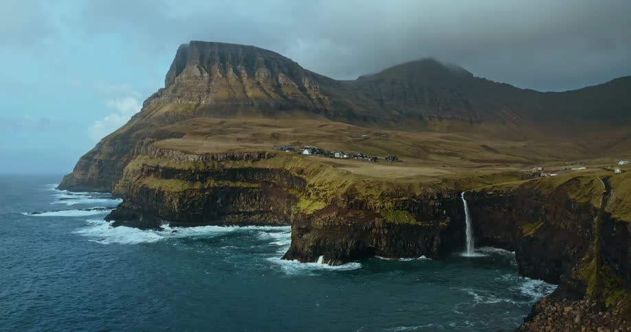 Waves crash against the island cliffs with a waterfall along the coastline as mountains stretch high into a hazy sky