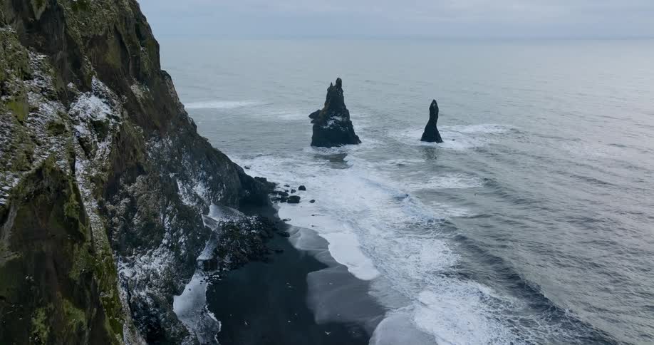 Rock formations stand tall in the water along the shoreline as seagulls fly over waves rolling in near a snow laced mountain