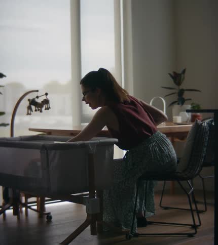 A woman is sitting at a table in front of windows with a laptop computer as she reaches into a playpen