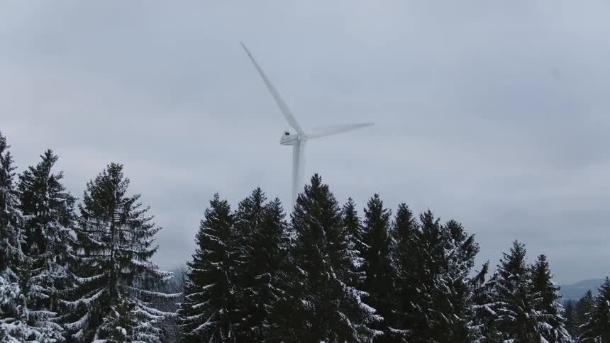 A wind turbine spins above snow covered trees in a forest