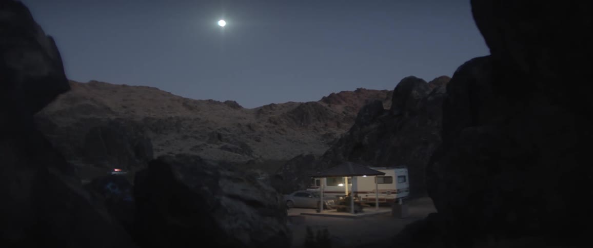 People sit under pavilion tent near rv and car parked in area surrounded by mountains under the moon